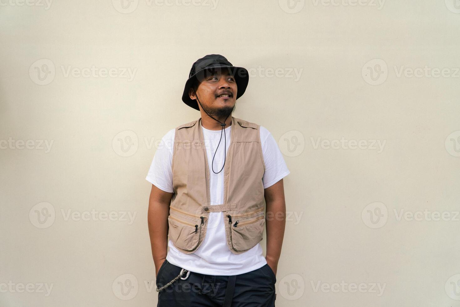 Macho mature man with beard and mustache wearing safari outfit isolated on beige background. Half body portrait of an adult Southeast Asian man posing wearing a vest, bucket hat and hip flask bottle photo