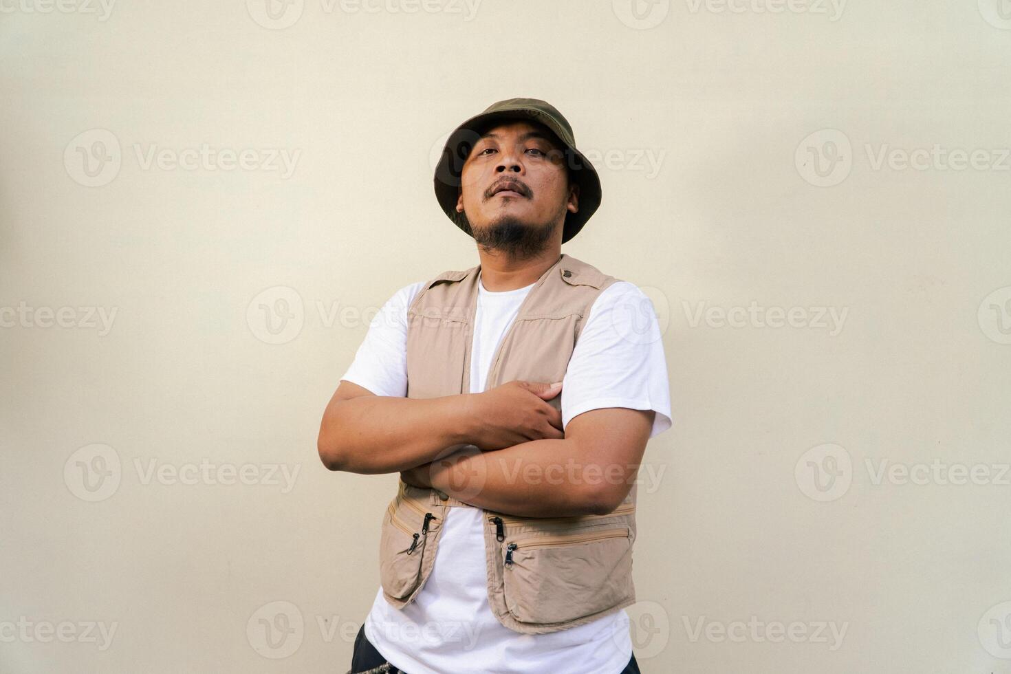 Macho mature man with beard and mustache wearing safari outfit isolated on beige background. Half body portrait of an adult Southeast Asian man posing wearing a vest, bucket hat and hip flask bottle photo