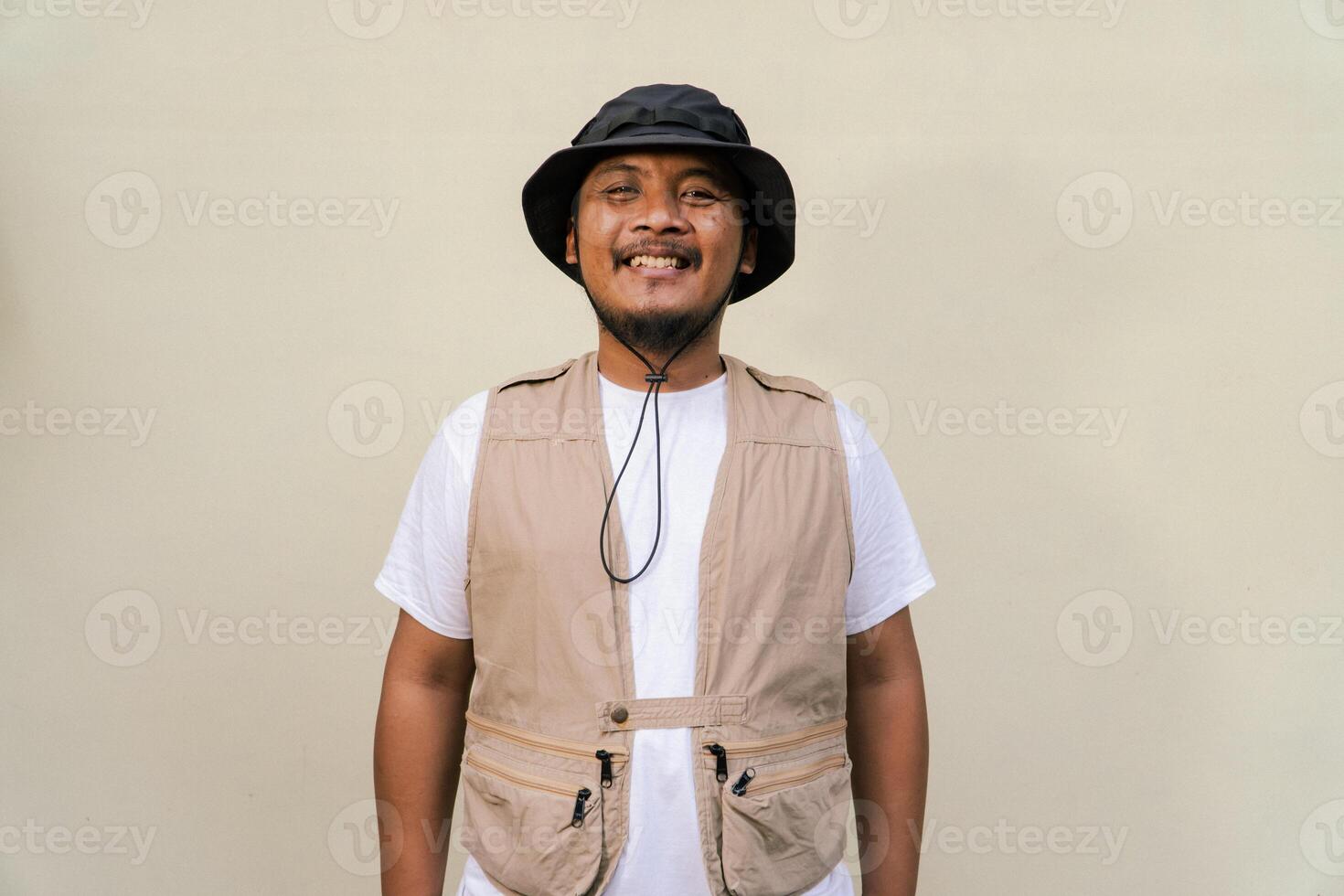 Macho mature man with beard and mustache wearing safari outfit isolated on beige background. Half body portrait of an adult Southeast Asian man posing wearing a vest, bucket hat and hip flask bottle photo