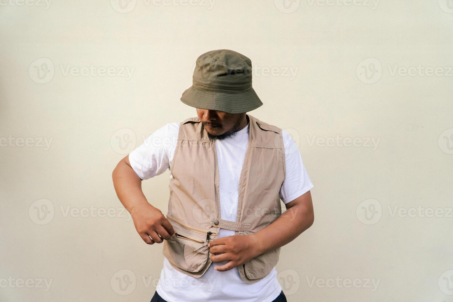 Macho mature man with beard and mustache wearing safari outfit isolated on beige background. Half body portrait of an adult Southeast Asian man posing wearing a vest, bucket hat and hip flask bottle photo