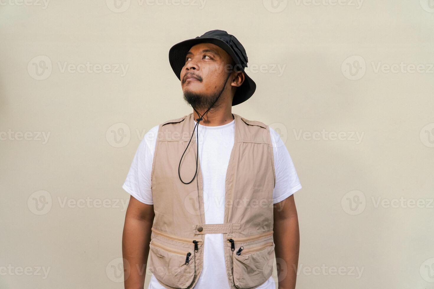 Macho mature man with beard and mustache wearing safari outfit isolated on beige background. Half body portrait of an adult Southeast Asian man posing wearing a vest, bucket hat and hip flask bottle photo