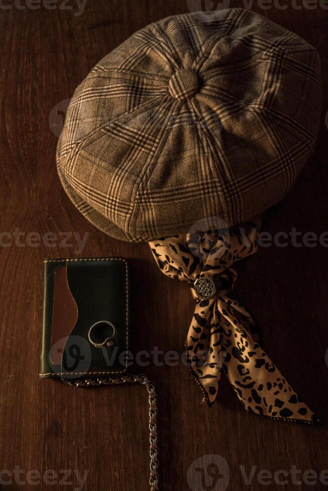 Classic newsboy cap or eight panel hat made of wool herringbone fabric on a wooden table in a dark room with various accessories around like watches, rings, handicrafts, chains and wallet photo