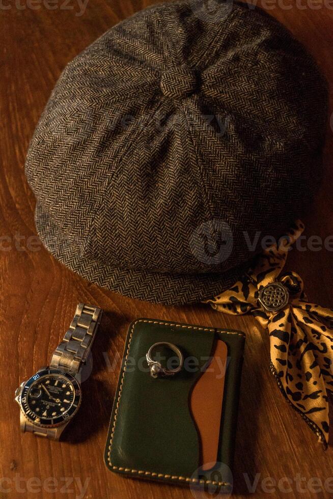 Classic newsboy cap or eight panel hat made of wool herringbone fabric on a wooden table in a dark room with various accessories around like watches, rings, handicrafts, chains and wallet photo