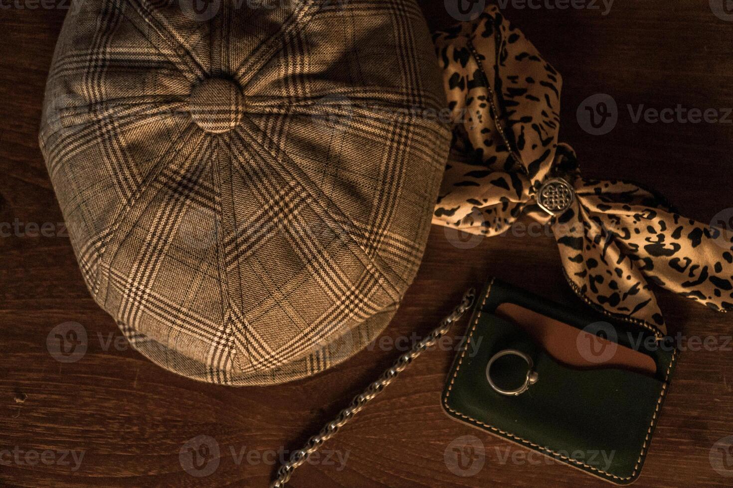 Classic newsboy cap or eight panel hat made of wool herringbone fabric on a wooden table in a dark room with various accessories around like watches, rings, handicrafts, chains and wallet photo