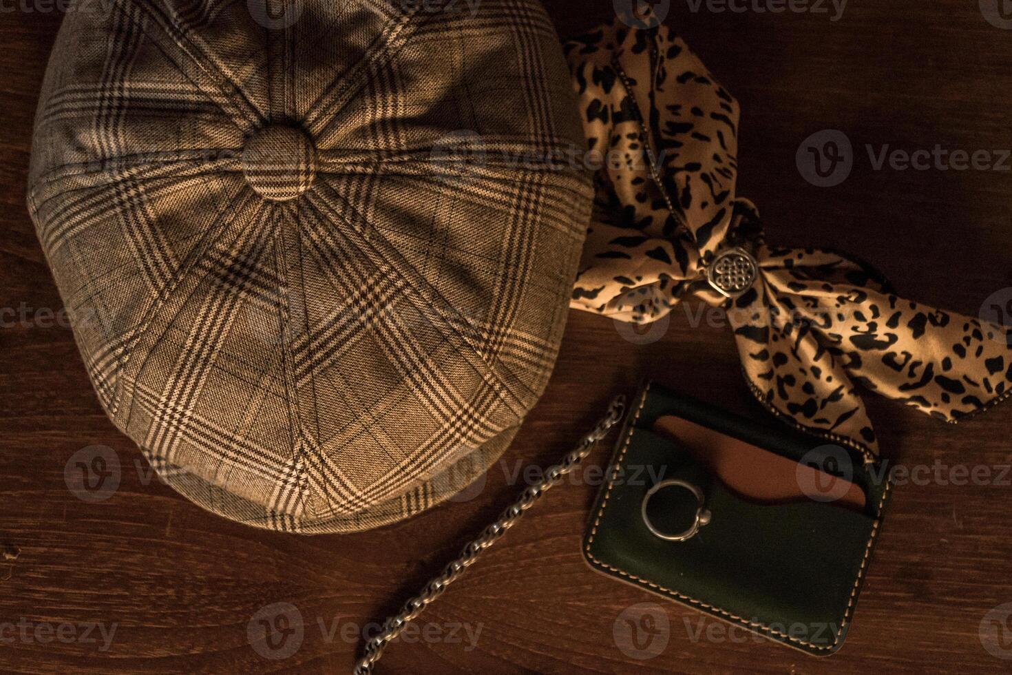 Classic newsboy cap or eight panel hat made of wool herringbone fabric on a wooden table in a dark room with various accessories around like watches, rings, handicrafts, chains and wallet photo