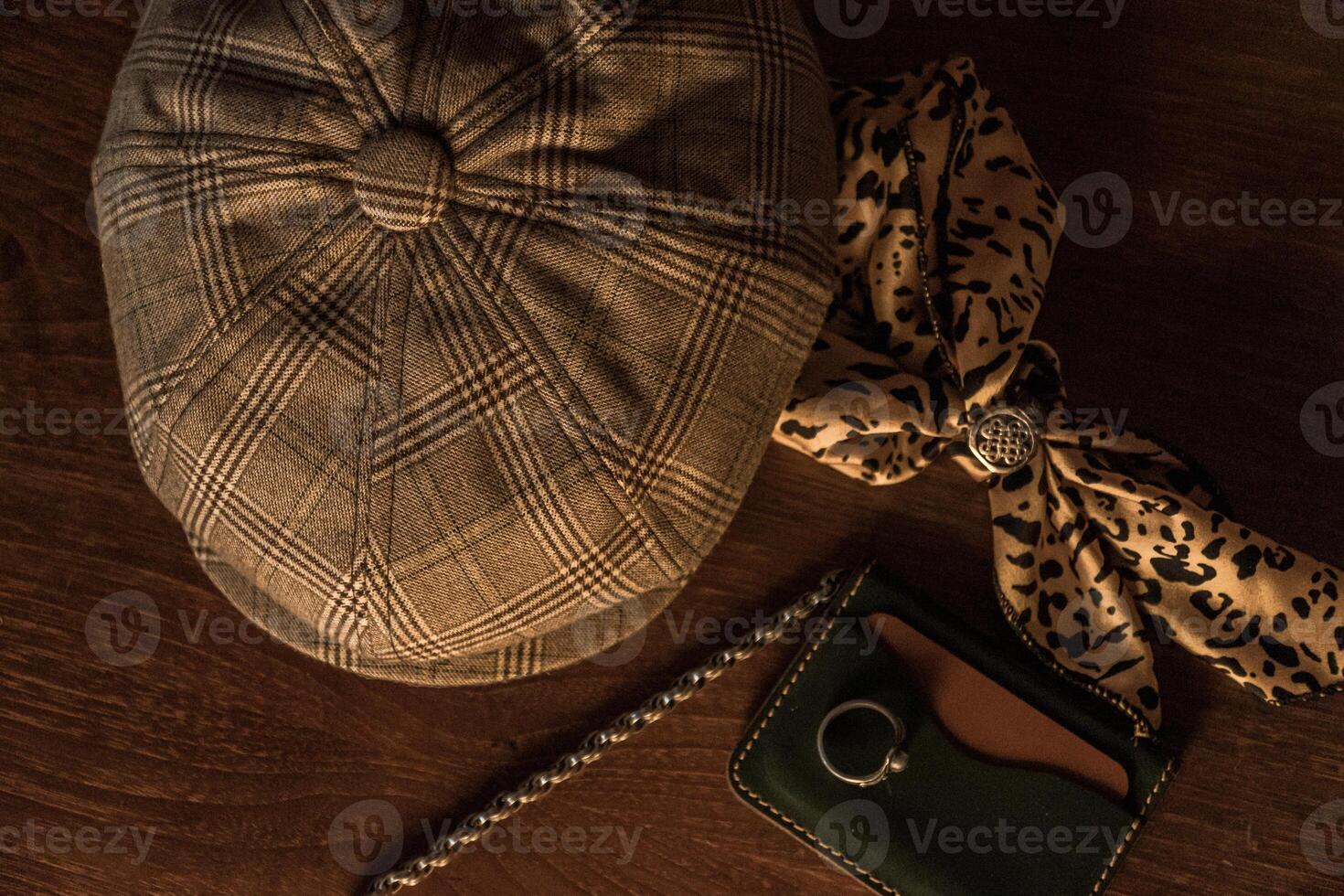 Classic newsboy cap or eight panel hat made of wool herringbone fabric on a wooden table in a dark room with various accessories around like watches, rings, handicrafts, chains and wallet photo