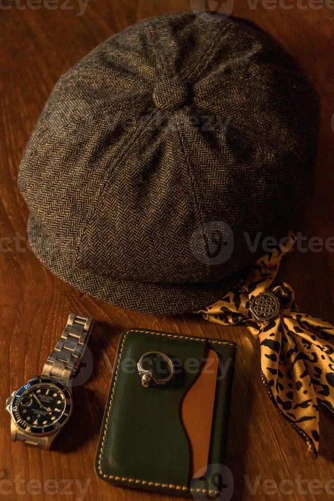 Classic newsboy cap or eight panel hat made of wool herringbone fabric on a wooden table in a dark room with various accessories around like watches, rings, handicrafts, chains and wallet photo