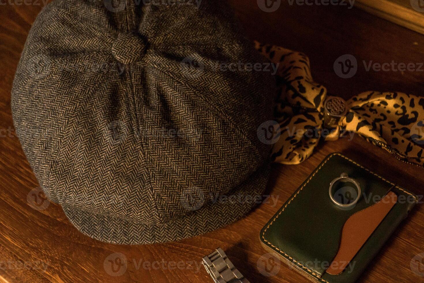 Classic newsboy cap or eight panel hat made of wool herringbone fabric on a wooden table in a dark room with various accessories around like watches, rings, handicrafts, chains and wallet photo