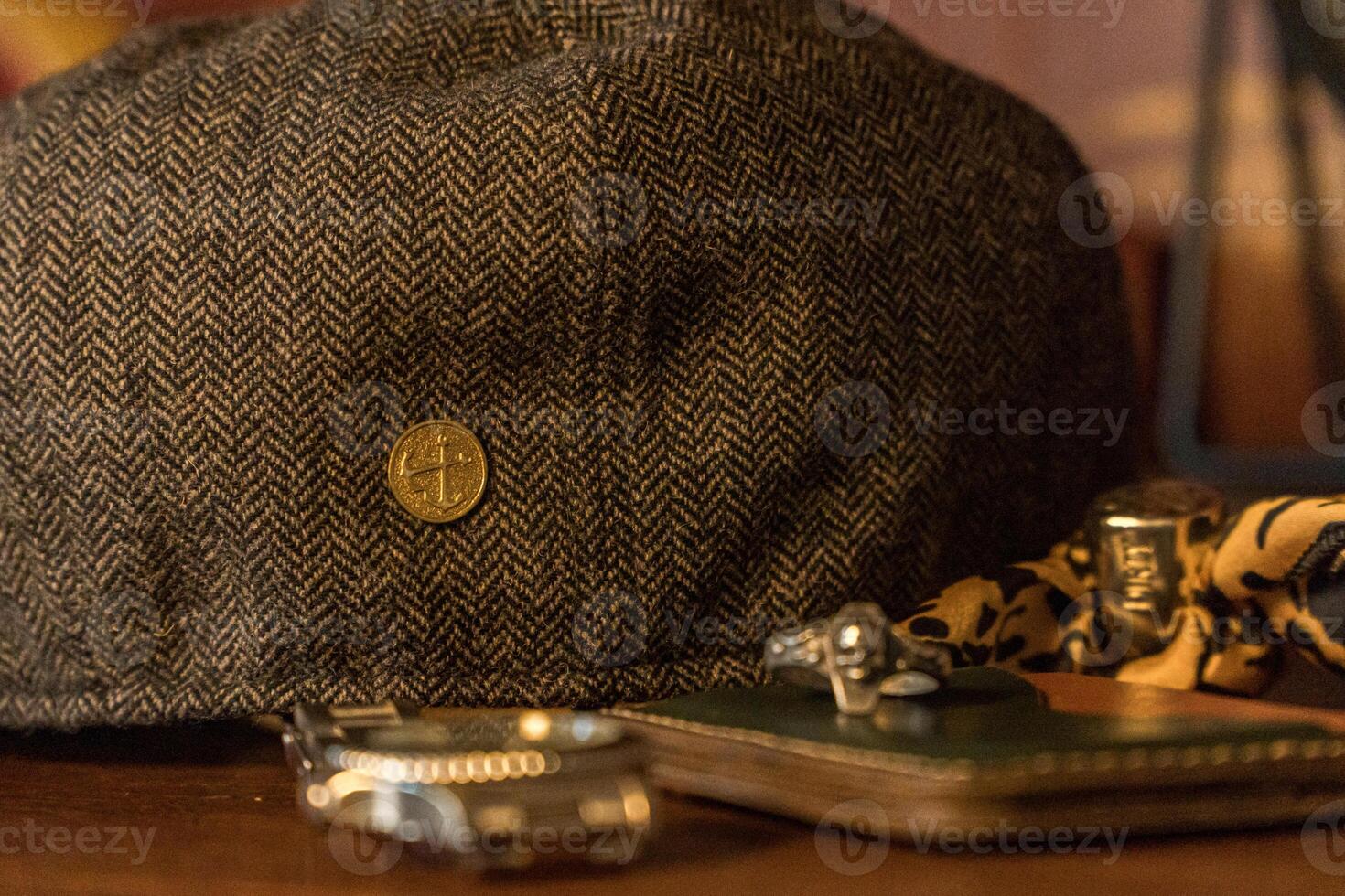 Classic newsboy cap or eight panel hat made of wool herringbone fabric on a wooden table in a dark room with various accessories around like watches, rings, handicrafts, chains and wallet photo