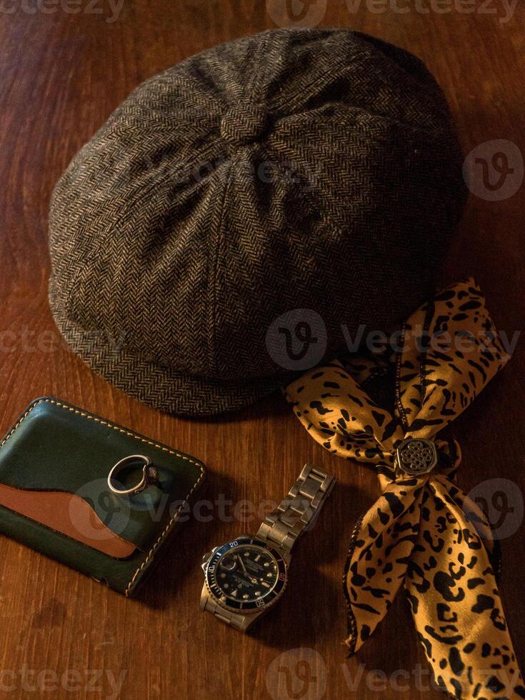Classic newsboy cap or eight panel hat made of wool herringbone fabric on a wooden table in a dark room with various accessories around like watches, rings, handicrafts, chains and wallet photo