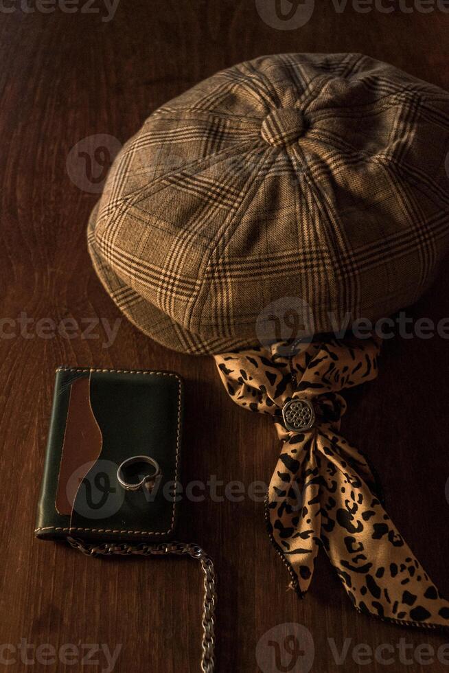 Classic newsboy cap or eight panel hat made of wool herringbone fabric on a wooden table in a dark room with various accessories around like watches, rings, handicrafts, chains and wallet photo