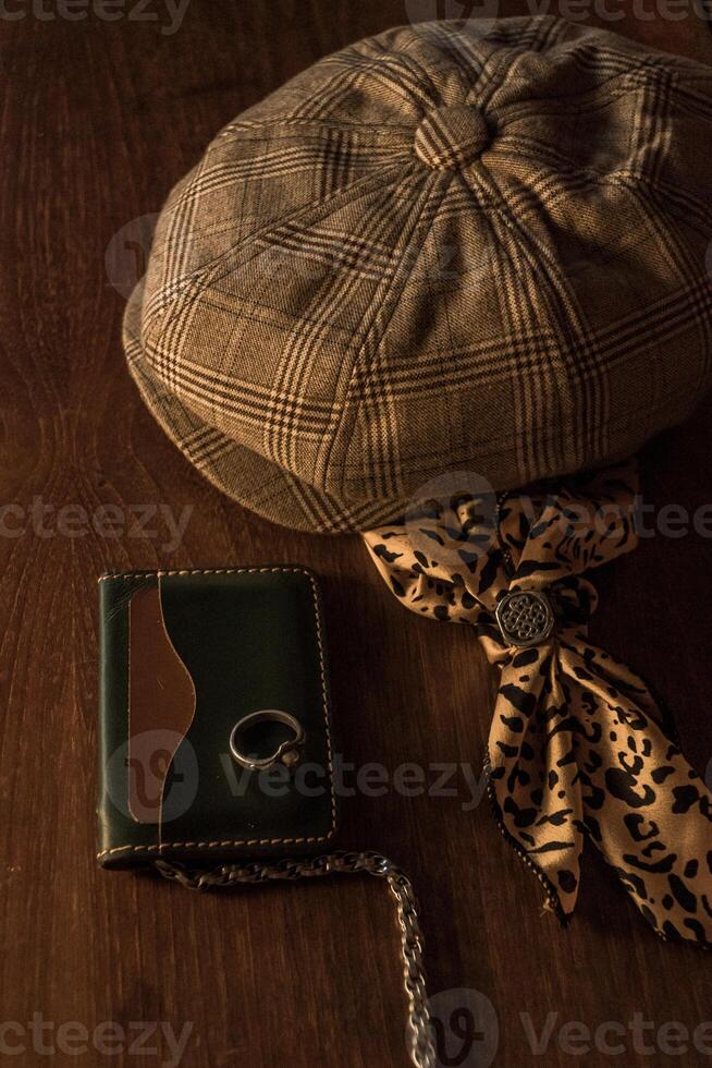 Classic newsboy cap or eight panel hat made of wool herringbone fabric on a wooden table in a dark room with various accessories around like watches, rings, handicrafts, chains and wallet photo