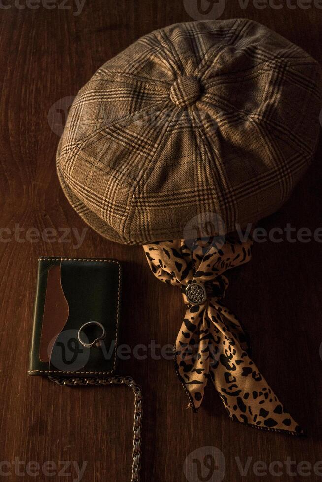 Classic newsboy cap or eight panel hat made of wool herringbone fabric on a wooden table in a dark room with various accessories around like watches, rings, handicrafts, chains and wallet photo