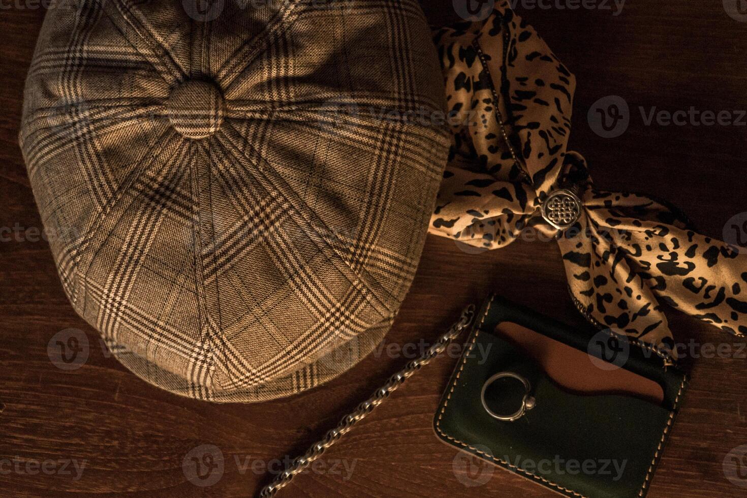 Classic newsboy cap or eight panel hat made of wool herringbone fabric on a wooden table in a dark room with various accessories around like watches, rings, handicrafts, chains and wallet photo