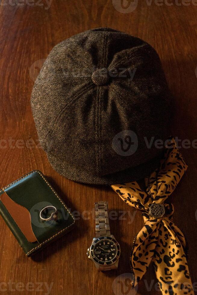 Classic newsboy cap or eight panel hat made of wool herringbone fabric on a wooden table in a dark room with various accessories around like watches, rings, handicrafts, chains and wallet photo