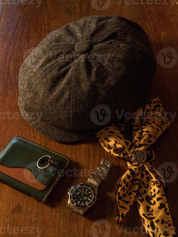 Classic newsboy cap or eight panel hat made of wool herringbone fabric on a wooden table in a dark room with various accessories around like watches, rings, handicrafts, chains and wallet photo