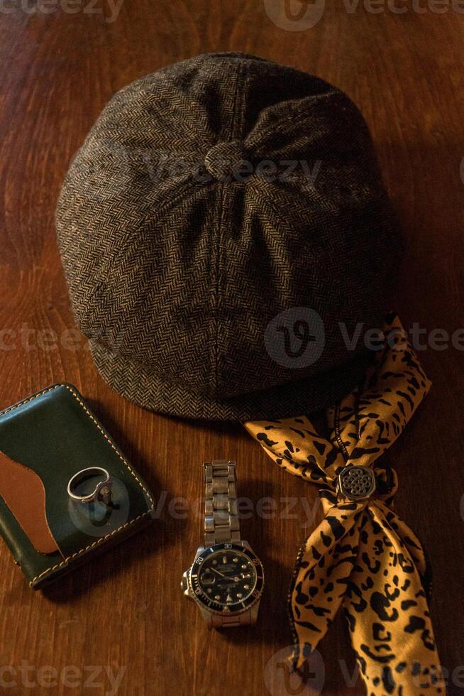 Classic newsboy cap or eight panel hat made of wool herringbone fabric on a wooden table in a dark room with various accessories around like watches, rings, handicrafts, chains and wallet photo