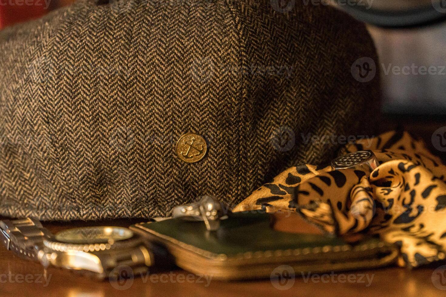 Classic newsboy cap or eight panel hat made of wool herringbone fabric on a wooden table in a dark room with various accessories around like watches, rings, handicrafts, chains and wallet photo