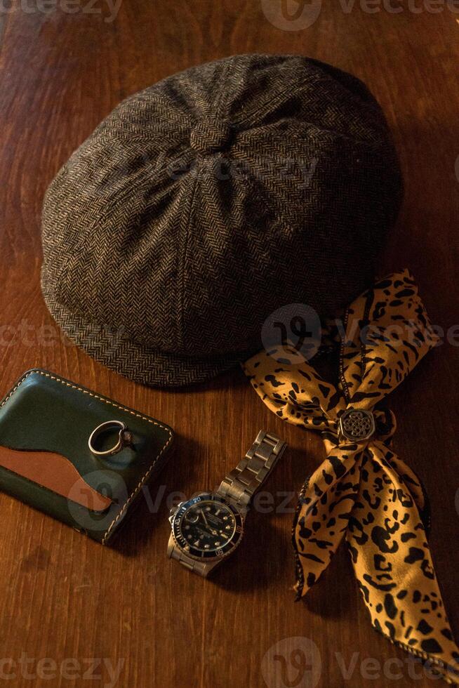 Classic newsboy cap or eight panel hat made of wool herringbone fabric on a wooden table in a dark room with various accessories around like watches, rings, handicrafts, chains and wallet photo