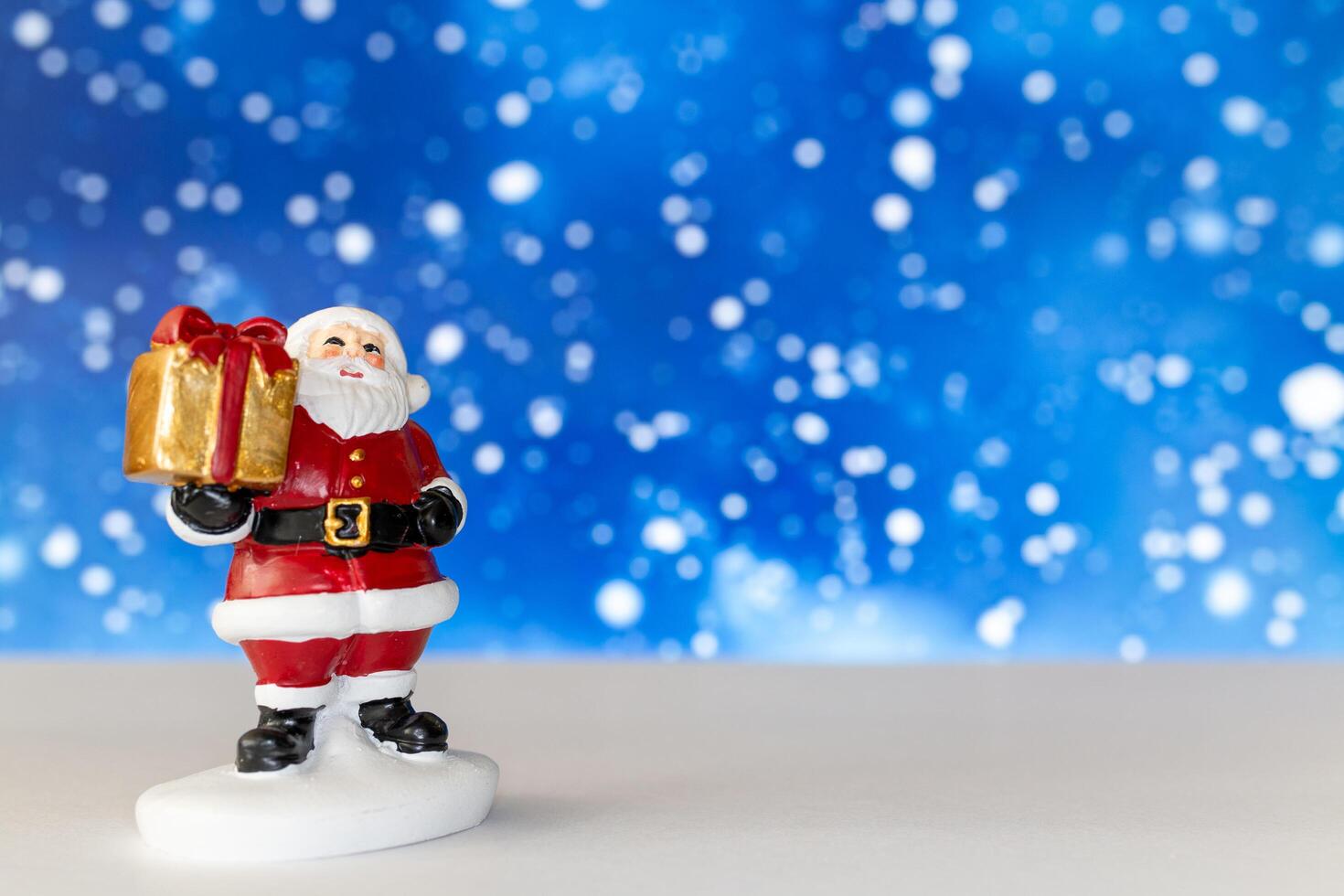 Cheerful Santa Claus figurine holding a golden gift box, set against a vibrant blue background with snowy bokeh effects photo