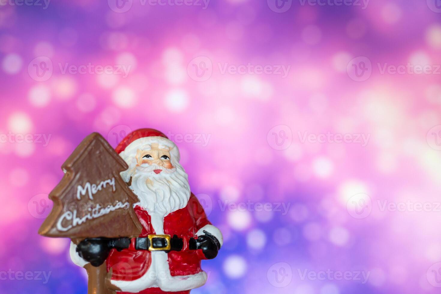Santa Claus figurine holding a Merry Christmas tree sign, set against a glowing bokeh background in warm festive tones photo