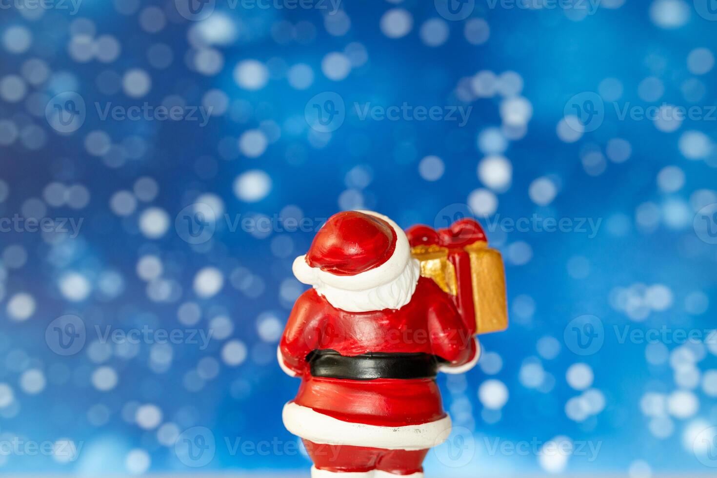 Cheerful Santa Claus figurine holding a golden gift box, set against a vibrant blue background with snowy bokeh effects photo
