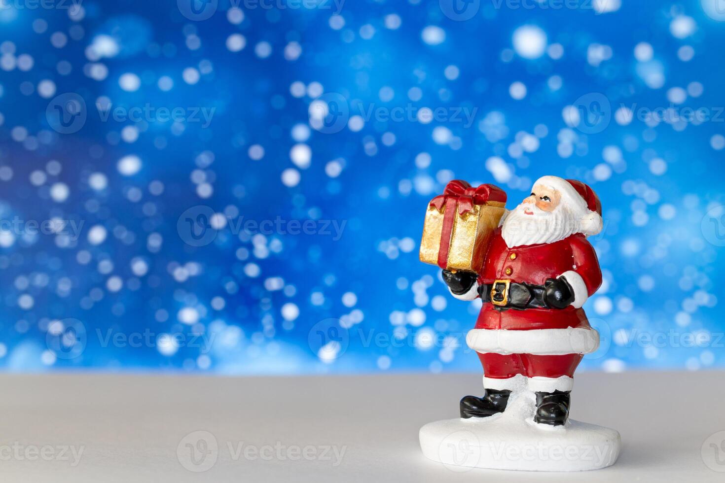 Cheerful Santa Claus figurine holding a golden gift box, set against a vibrant blue background with snowy bokeh effects photo