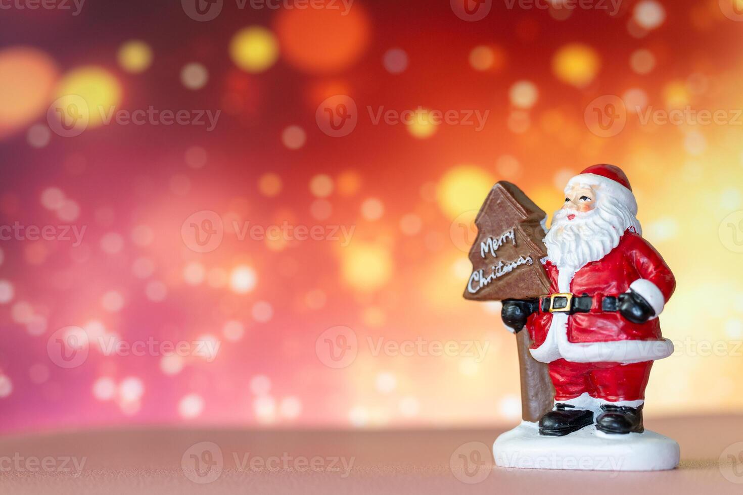 Santa Claus figurine holding a Merry Christmas tree sign, set against a glowing bokeh background in warm festive tones photo