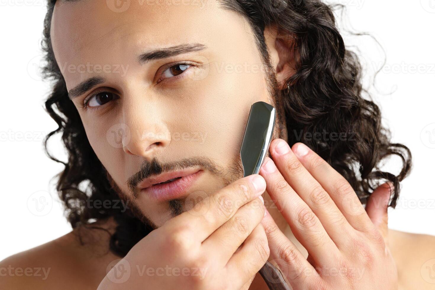 Man with curly hair grooming his beard carefully using a straight razor against a plain white background photo