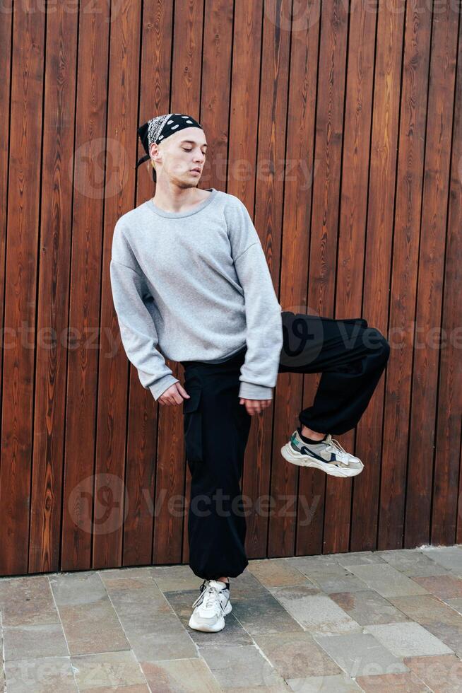 A young man dances in front of a wood panel wall, wearing a grey sweater, black pants, and white sneakers photo