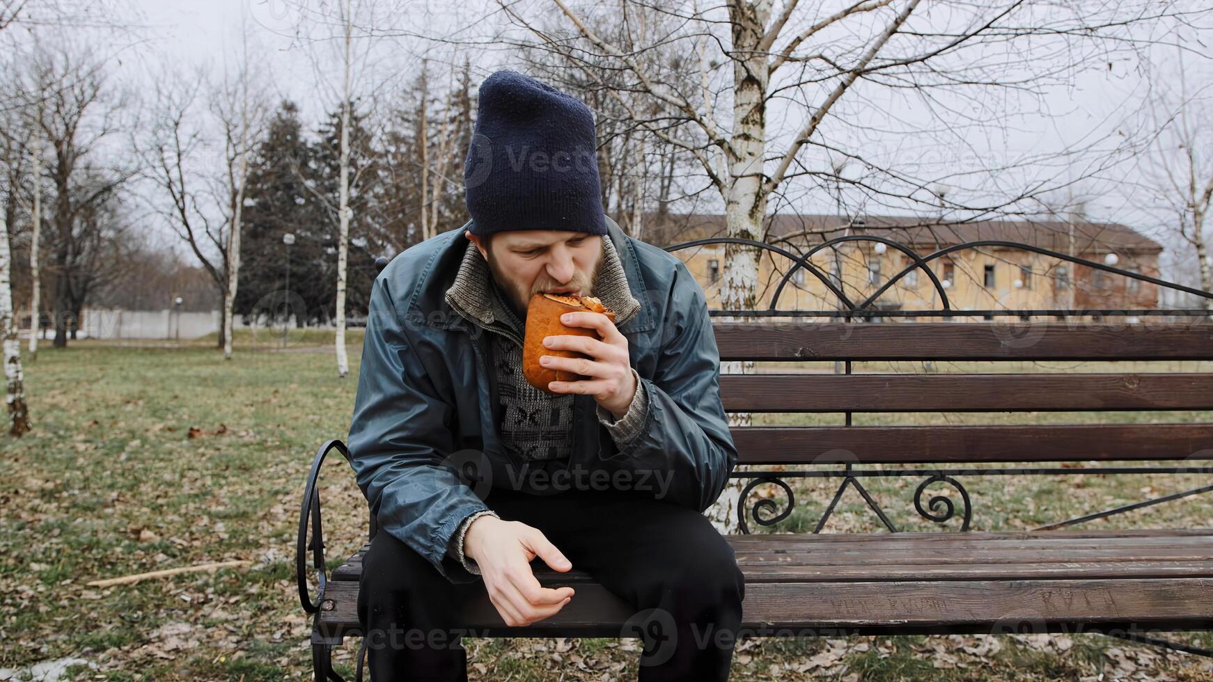 Hungry bearded homeless man sits on a bench and eats a bun in a city park. Below poverty line. Male tramp in dirty clothes with cap and hat. Beggar. Illegal immigrant. Drunkenness, alcoholism photo