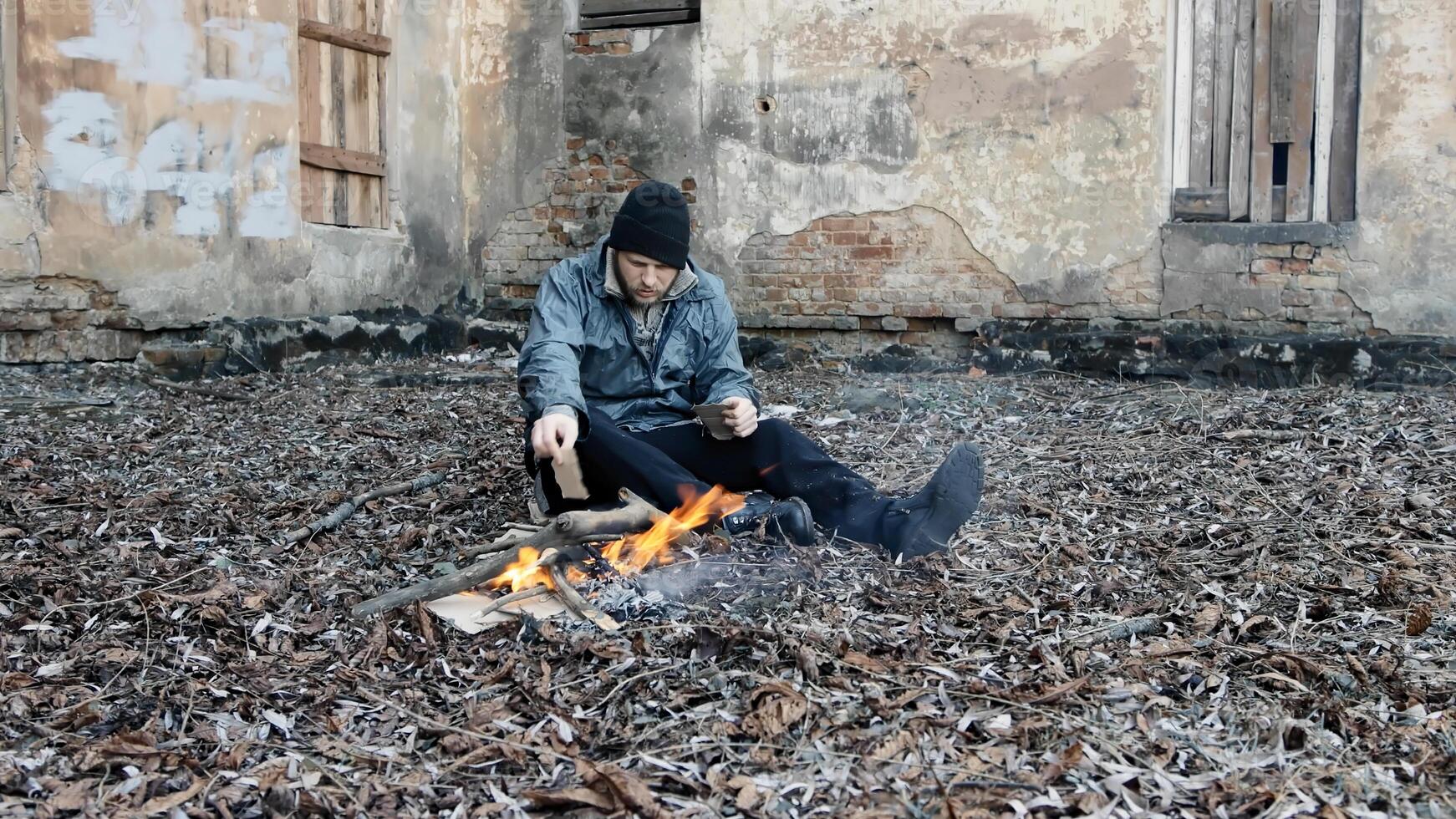 Homeless man sits on ground and pushes pieces of cardboard into fire to keep warm from cold. Tramp in dirty clothes and hat cap. Below poverty line. Illegal immigrant. Male refugee. Fugitive beggar photo