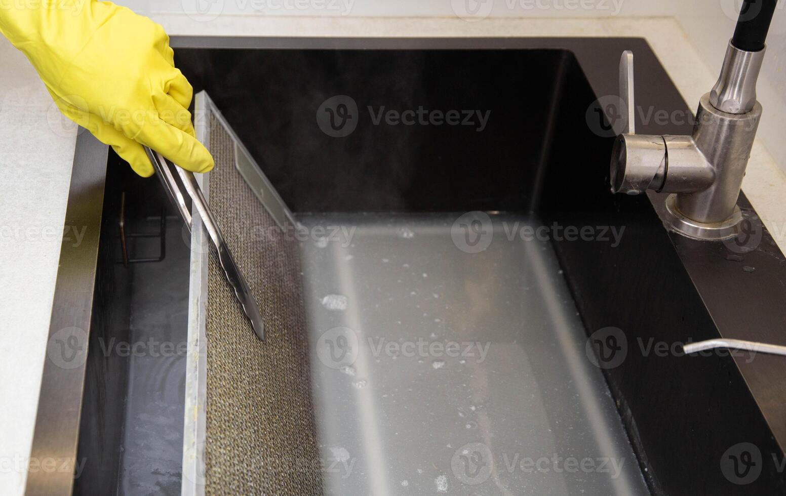 A girl's hand in a yellow glove turns over a filter from a kitchen hood with tongs in the cleaning mixture of an oxygen purifier for the kitchen. Chemical reaction photo