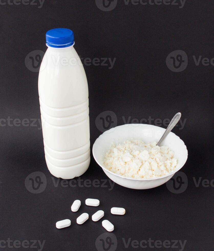 A bottle of milk and a plate of cottage cheese on a black background near lie pills of antibiotics, close-up, pills photo