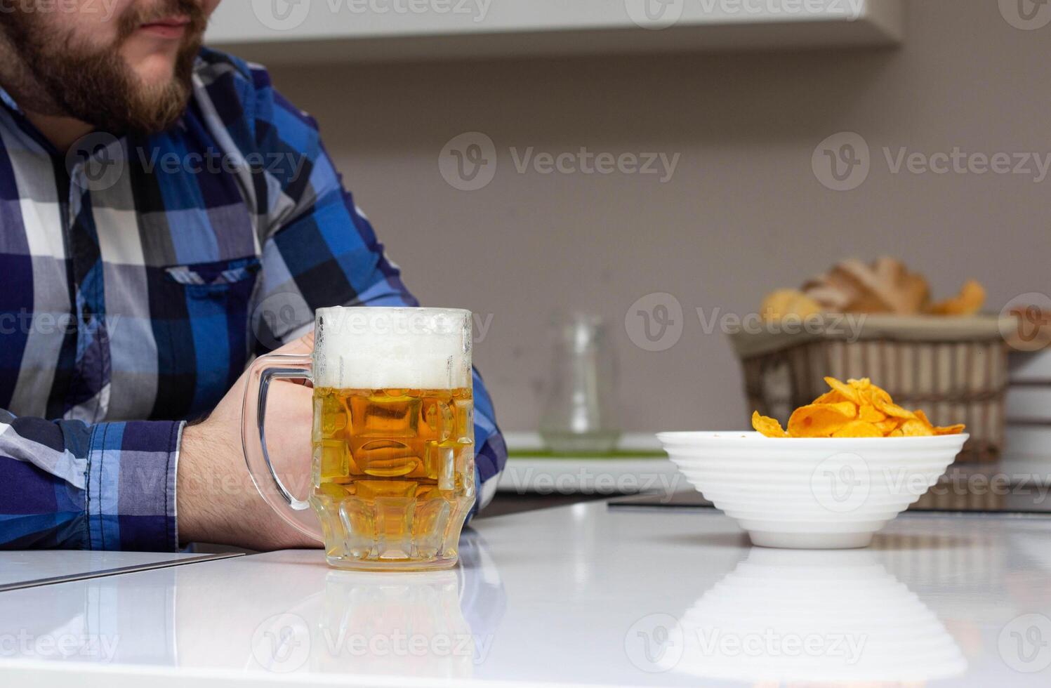A man with a beard is sitting in the kitchen, on the table is a glass of beer and chips, junk food, domestic photo