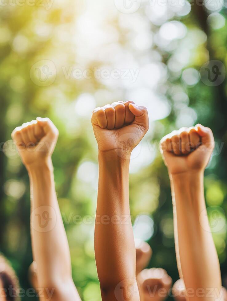 A diverse crowd of individuals raises their fists high in a display of unity, symbolizing solidarity among all attendees during a peaceful protest taking place in a beautifully sunlit park setting photo