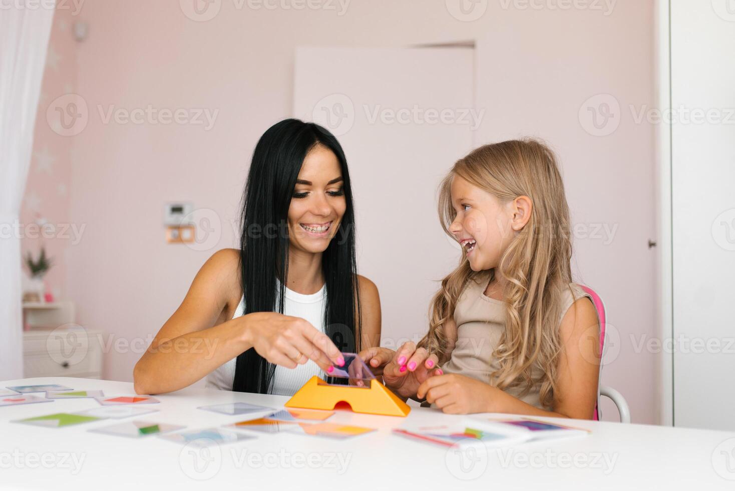 Smiling mother and a little daughter play a logical strategy board game together at the table in the nursery, a happy family is engaged in logical activities at home photo