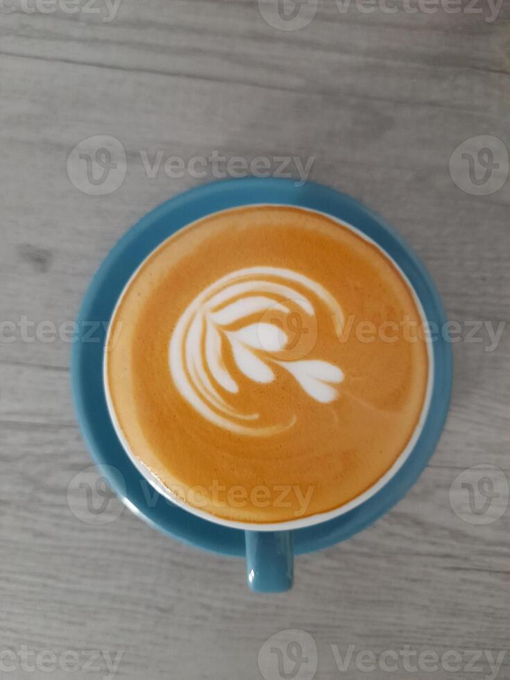a cup of latte placed on a blue saucer, sitting on a wooden table. The latte has a delicate latte art design on its frothy surface, resembling a stylized flower or leaf. photo