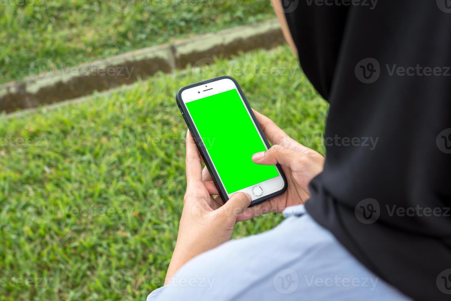 A joyful woman in a black hijab and blue shirt smiles brightly while holding a smartphone, standing at the edge of a beautiful park sidewalk photo