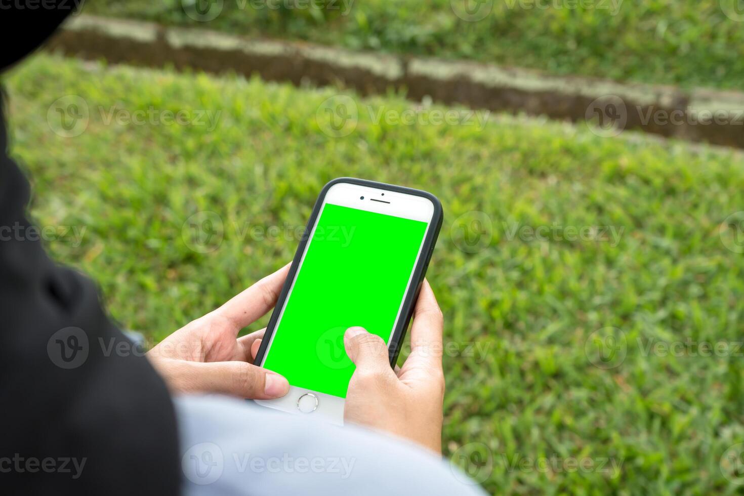 A joyful woman in a black hijab and blue shirt smiles brightly while holding a smartphone, standing at the edge of a beautiful park sidewalk photo