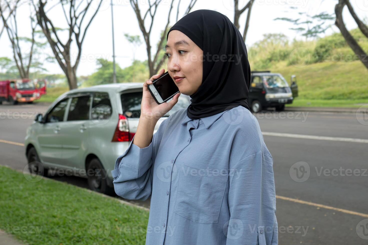A woman wearing a black hijab and blue shirt smiles while on a call, occasionally looking at the camera. She appears cheerful and engaged, exuding a positive, friendly vibe photo