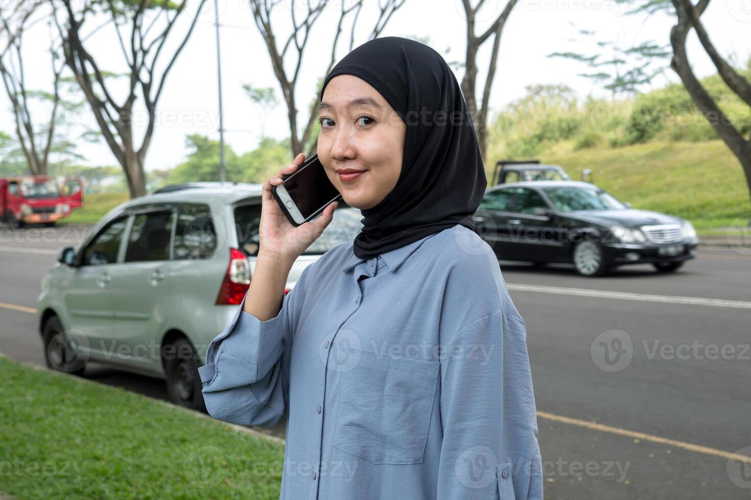 A woman wearing a black hijab and blue shirt smiles while on a call, occasionally looking at the camera. She appears cheerful and engaged, exuding a positive, friendly vibe photo