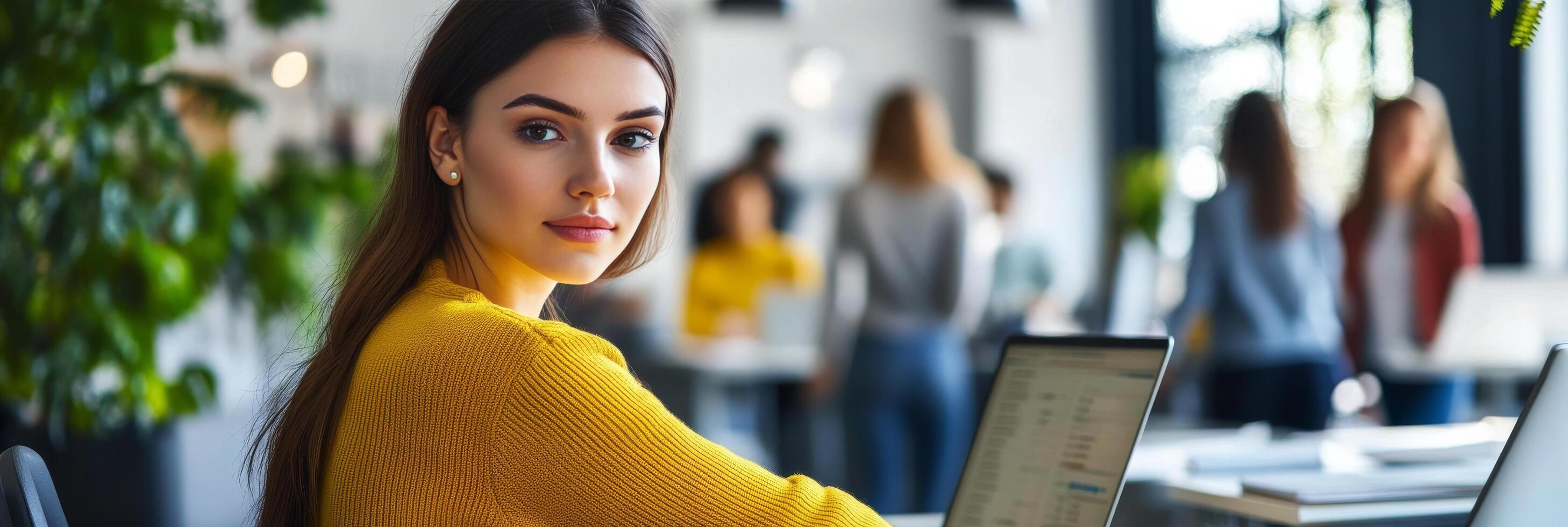 Young woman focused on laptop in modern workspace during daytime with colleagues in background photo