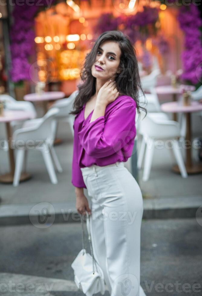 Young beautiful brunette woman in a pink jacket and white pants posing on the street. Portrait of a beautiful young woman in a purple blouse and white pants posing in an outdoor cafe on the street photo