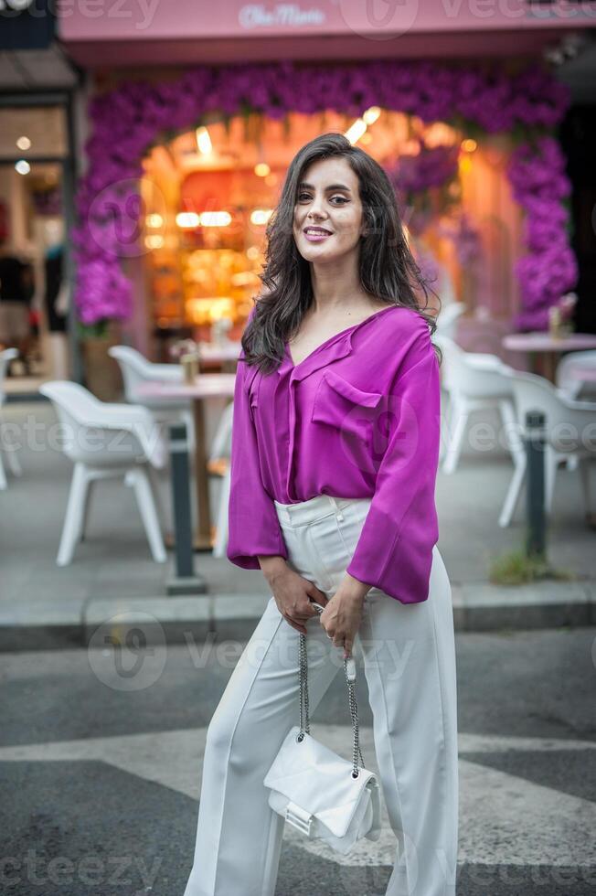 Young beautiful brunette woman in a pink jacket and white pants posing on the street. Portrait of a beautiful young woman in a purple blouse and white pants posing in an outdoor cafe on the street photo