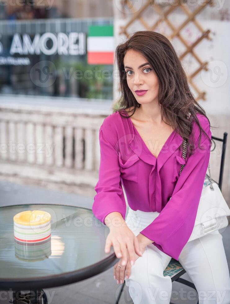 Young beautiful brunette woman in a pink jacket and white pants posing on the street. Portrait of a beautiful young woman in a purple blouse and white pants posing in an outdoor cafe on the street photo