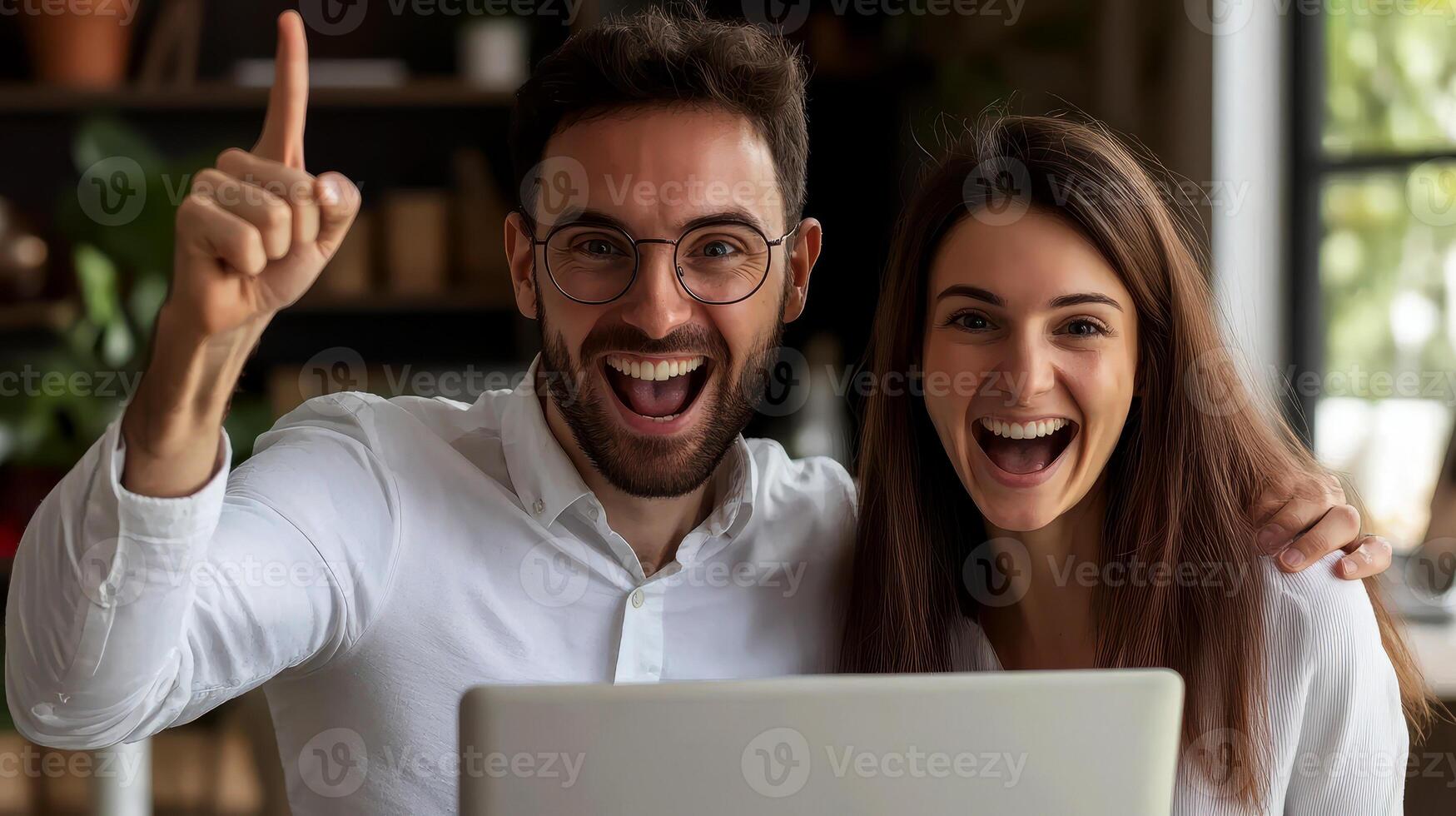 A man and a woman are sitting at a desk with a computer monitor in front of them. The man is smiling and holding his hands up in the air, while the woman is also smiling. Scene is happy and positive. photo