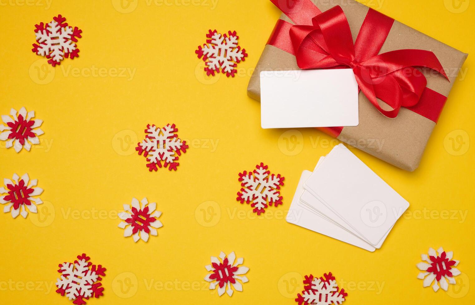 Christmas gift wrapped in red ribbon on yellow background, stack of white paper business cards. Top view photo