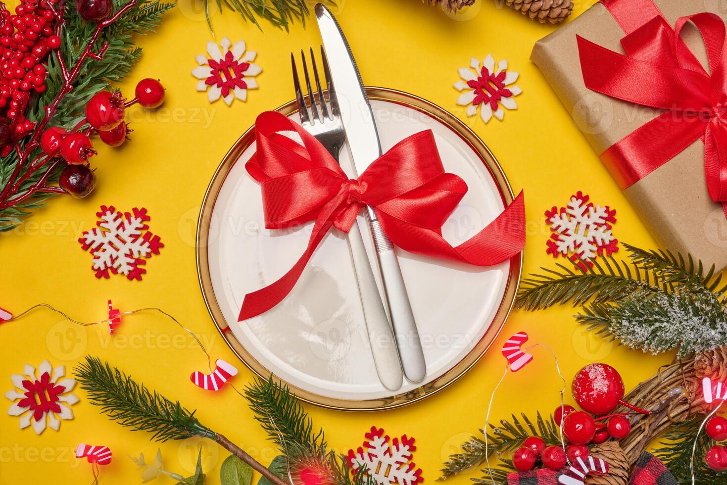 Festive composition with a plate and cutlery tied with a red ribbon, a gift, fir branches, pine cones and decorative snowflakes on a bright yellow background photo