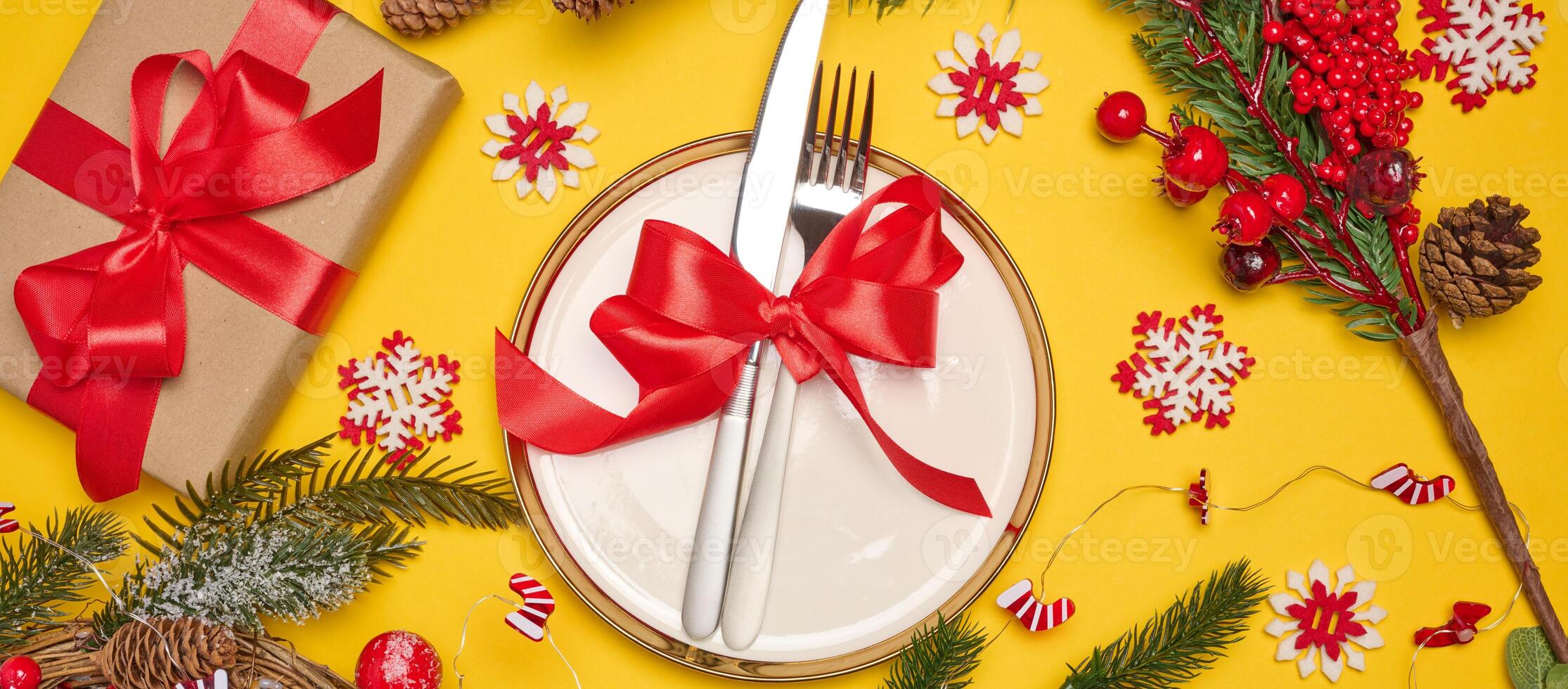 Festive composition with a plate and cutlery tied with a red ribbon, a gift, fir branches, pine cones and decorative snowflakes on a bright yellow background photo